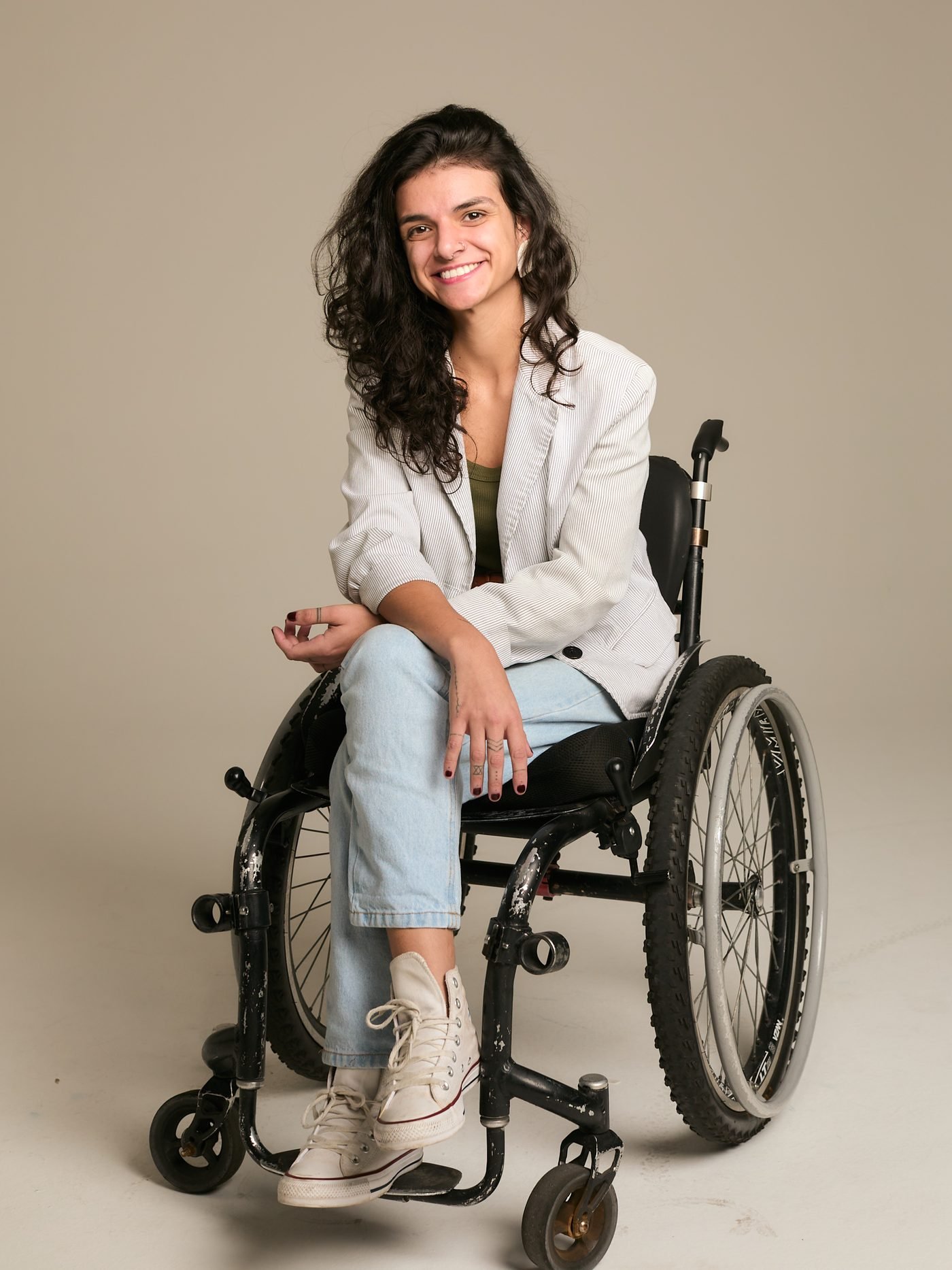 Retrato de Talita Cadeirante em estúdio, de blazer claro e calça jeans, sentada em sua cadeira de rodas
